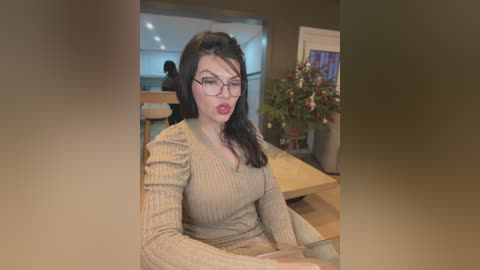 Media: Video of a fair-skinned woman with long black hair and glasses, wearing a beige ribbed sweater, sitting indoors near a wooden table with a potted plant. Background includes a blurry view of a modern kitchen.