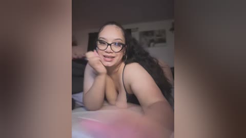 Video of a smiling, voluptuous woman with long black hair, wearing glasses, lying on a bed in a dimly lit bedroom, with a blurred background.