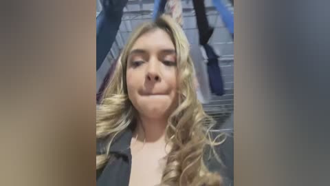 Video of a blonde woman with wavy hair and closed eyes, smiling slightly. She's wearing a black top, standing in front of a blurred background featuring a staircase and blue objects.