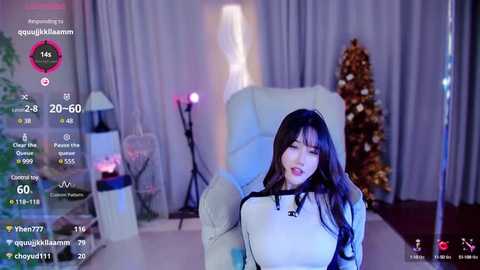 Media: Video of an Asian woman with long black hair in a white sweater, seated in a dimly-lit living room with a Christmas tree and a white chair, displaying a live stream.