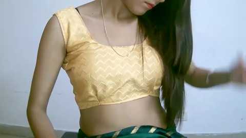 Media: Video of a young woman with long dark hair, wearing a gold sequin crop top and green skirt, against a plain white wall background.