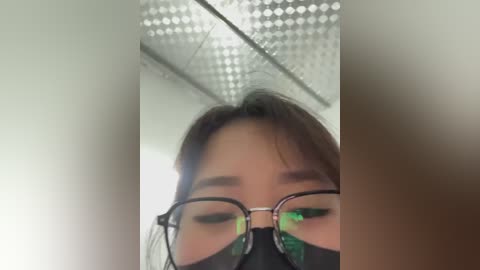 Media: Video of a close-up of an Asian woman with glasses and a black mask, capturing her partially obscured face in a fluorescent-lit room with a ceiling grid.