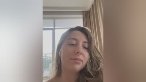 Video of a young Caucasian woman with fair skin and wavy, shoulder-length blonde hair, wearing minimal makeup, standing indoors with a serene expression, near a window with beige curtains and a balcony view.