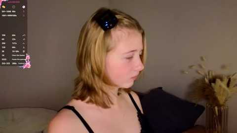 Media: Video of a fair-skinned, blonde-haired young woman with a blue hair clip, wearing a black bra, sitting indoors, looking to the right. Background includes a gray wall, a vase with dried flowers, and a black pillow.