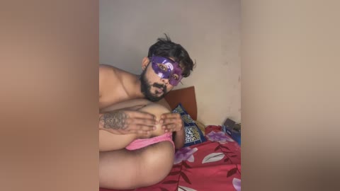 Media: Video of a topless, dark-skinned man with a purple mask and beard, holding his large, exposed breasts in a messy bedroom.