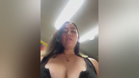 Media: Video of a plus-size Latina woman with long dark hair, wearing a black lace bra, standing in a dimly lit room with fluorescent lights. She has a neutral expression and medium skin tone.