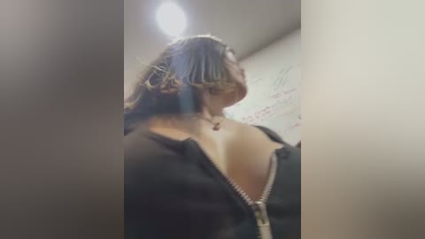 Media: Video of a woman with shoulder-length, curly hair, wearing a black, unzipped jacket, standing in a dimly lit room with white walls and a visible ceiling light.