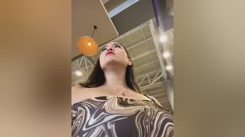 Media: Video of a woman with long brown hair, wearing a strapless top with a black and white pattern, looking up at a ceiling with a round, orange light fixture.