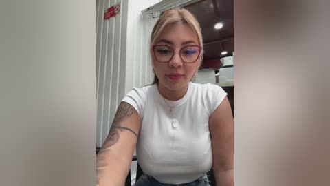 Media: Video of a young woman with fair skin, light blonde hair, and round glasses, wearing a white, short-sleeved, ribbed top, sitting in an indoor, modern setting with a large window and visible ceiling lights.