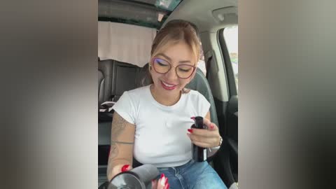 Media: Video of a smiling, light-skinned woman with blonde hair in glasses, wearing a white t-shirt, blue jeans, and a black wristwatch, texting on a smartphone in the backseat of a car.