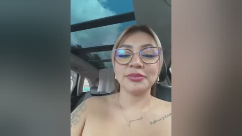 Media: Video of a topless woman with light skin, wearing glasses, sitting in a car with a blue sky and clouds visible through the windshield.