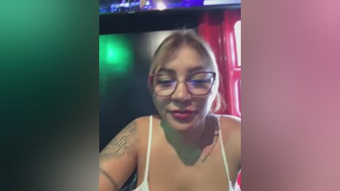 Media: Video of a light-skinned, smiling woman with glasses, tattoos on her arms, wearing a white tank top, in a dimly lit room with red and green lights.