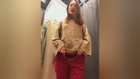 Media: Video of a young woman with light skin and long brown hair, wearing a beige blouse and red pants, standing in a fitting room with beige walls and a gray curtain.