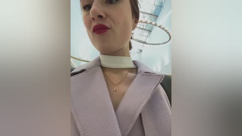 Media: Video of a woman with fair skin, brown hair, and red lipstick, wearing a light purple coat and white choker necklace, against a blurred, modern indoor background with a circular ceiling light.