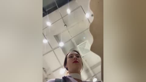 Media: Video of a woman in a white shirt and red lipstick, looking up at a ceiling with bright lights and grid pattern, taken from below.