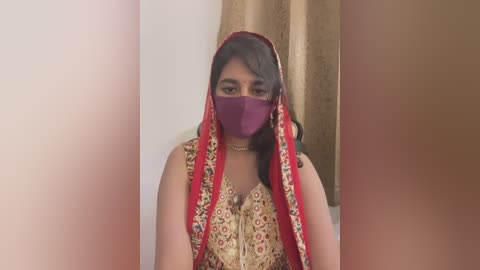 Media: Video of a woman with medium brown skin wearing a red and gold sari with intricate embroidery, a purple mask covering her nose and mouth, and a red veil. Background features beige curtains and a white wall.