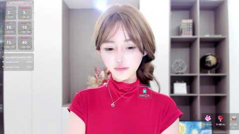 Media: Video of a young Asian woman with light skin and straight brown hair, wearing a red turtleneck, standing in a modern office with white walls and shelves filled with books and decor.