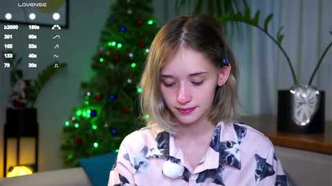 Media: Video of a young Caucasian woman with shoulder-length blonde hair, wearing a white shirt with black dog pattern, smiling, in a cozy, festive living room with a decorated Christmas tree, greenery, and potted plants.