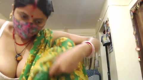 Media: Video of an Indian woman in a green sari, showing cleavage, adjusting her sari in a cluttered, dimly lit room with clothes hanging on the wall.