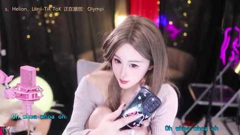 Media: Video of a slender, light-skinned Asian woman with long brown hair, wearing a beige knitted sweater, sitting on a chair. She is holding a pink phone, looking at it with a slightly confused expression. Background features a dimly lit room with blurred lights.