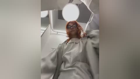 Media: Video of a person with red hair, wearing glasses and a white lab coat, standing inside a sterile, brightly lit lab with circular lights.