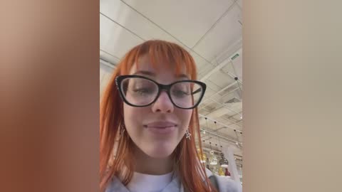 Media: A video of a young woman with long, fiery red hair, wearing black-rimmed glasses and a white top, standing in a brightly lit, modern indoor space with white ceiling panels and fluorescent lighting.