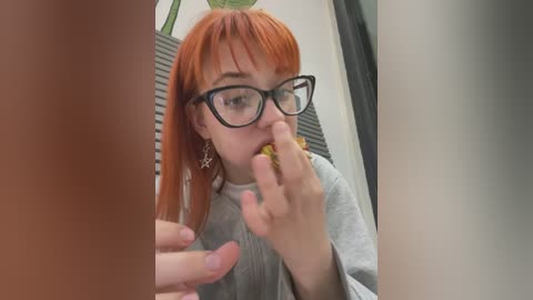 Media: Video of a young woman with straight, shoulder-length orange hair, wearing black-rimmed glasses, grey top, and eating a sandwich. Background includes a window, potted plant, and a blurred person.