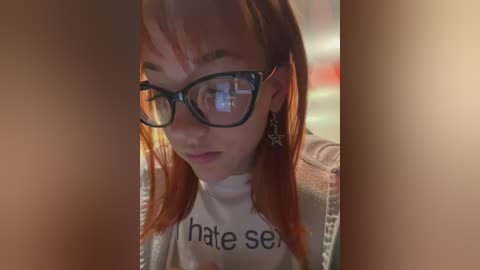 Media: Video of a young woman with long red hair, wearing black-rimmed glasses and a white t-shirt with \"I hate sex\" written in black. She has a neutral expression. The background is blurred, creating a soft focus effect.