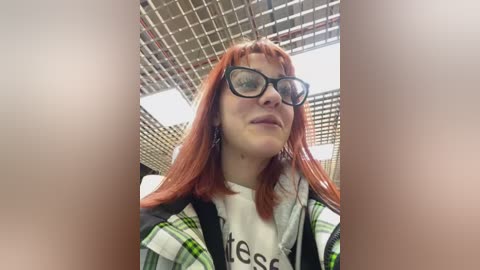 Media: Video of a young woman with straight, shoulder-length red hair, wearing black-rimmed glasses and a green and white plaid shirt, standing indoors under a grid-patterned ceiling.