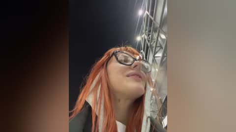 Media: Video of a young woman with long, straight, orange hair, wearing black-rimmed glasses, looking upward into a bright spotlight, possibly on stage. She has fair skin and a slight smile.