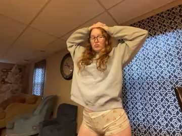 Media: A video of a young Caucasian woman with long, wavy red hair, wearing glasses, a gray sweatshirt, and patterned pajama shorts, stretching in a cozy living room with patterned wallpaper and a clock on the wall.