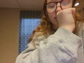 Media: Video of a fair-skinned woman with long, wavy red hair, wearing glasses, a gray sweater, and a contemplative expression, leaning on a table, against a patterned wall.