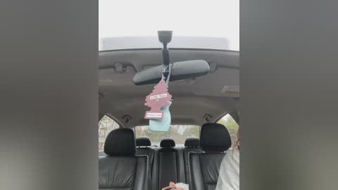 Media: Video of a car interior showing a person sitting in the back seat. The car has black leather seats, a light blue air freshener dangling from the rearview mirror, and a partially visible person wearing a white shirt.