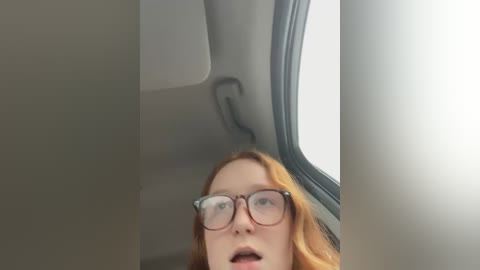 Media: Video of a young woman with long red hair, wearing glasses, looking out a car window with a reflective surface.