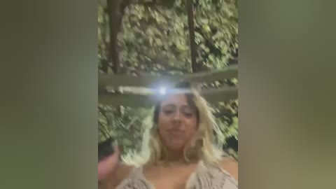 Media: A video captures a blonde woman with shoulder-length hair and a light-colored sleeveless top, standing in a dense forest. She holds a camera, taking a photo, with blurred greenery in the background.