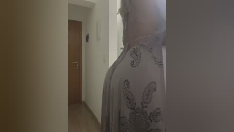 Media: A video of a hallway with a person wearing a white dress adorned with black paisley patterns, standing in the foreground. The background reveals a wooden door and beige walls, creating a dimly lit, serene atmosphere.