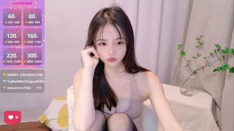 Media: A young Asian woman with long black hair sits on a bed, wearing a sleeveless top and black stockings. The room has a minimalist decor with beige curtains and a green plant.