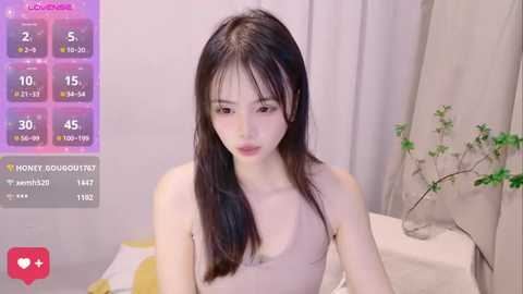 Media: A video of a young East Asian woman with long black hair, fair skin, and a slim physique, wearing a beige tank top, seated indoors.