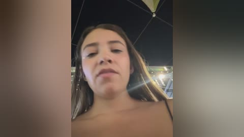 Media: Video of a young woman with light skin and long brown hair, wearing a black top, under an open black umbrella, taken from a low angle, showing a clear, focused facial expression.