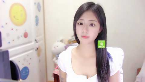 Media: Video of a young Asian woman with long black hair, wearing a white blouse, standing in a colorful, playful room with decorative wall stickers.