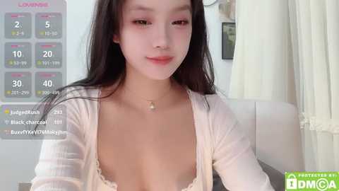 Media: Video of a young Asian woman with long black hair, wearing a white cardigan, revealing a lacy bra, sitting indoors.