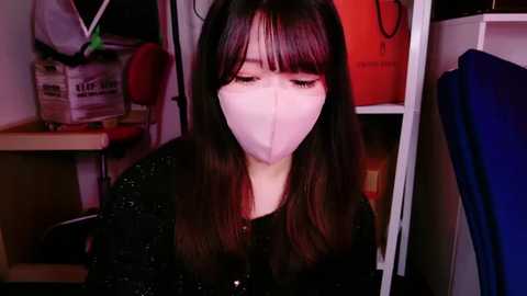 Media: Video of a young Asian woman with long, straight black hair, wearing a white surgical mask, seated in a dimly lit room, surrounded by clutter.