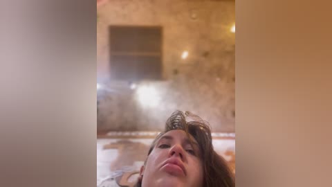 Media: Video of a woman with wet, tousled brown hair, eyes closed, lying in a steaming hot tub, surrounded by blurred, beige stone walls and dim lighting, creating a relaxed, steamy atmosphere.