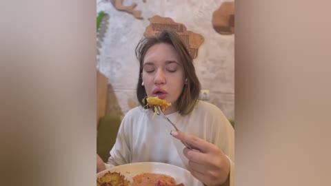 Media: A video of a young woman with straight, shoulder-length brown hair, wearing a white long-sleeve shirt, eating spaghetti in a cozy, indoor setting with a rustic, stone wall and wooden structures in the background.