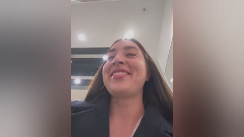 Media: Video of a smiling Latina woman with long brown hair, wearing a black shirt, taken from a low angle, indoors with bright lights and blurred background.