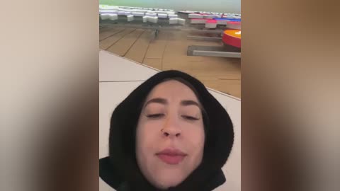 Video of a woman with light skin, closed eyes, and a black hijab, lying on a wooden floor with colorful, empty bumper cars in the background.