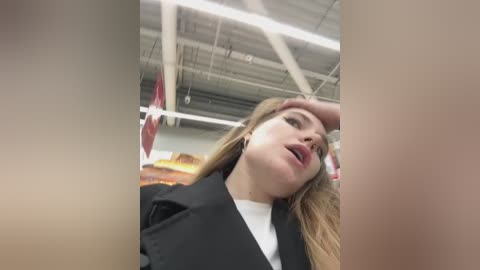 Media: Video of a young Caucasian woman with light skin and long blonde hair, wearing a black blazer over a white shirt, appearing distressed, with a hand on her forehead. Background features a supermarket with fluorescent lights, red signs, and a warm, colorful painting.