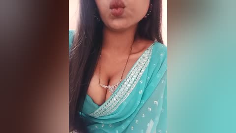 Media: Video of a South Asian woman with long dark hair, wearing a turquoise sari with white embroidery, making a pouty kiss, set against a teal and brown background.