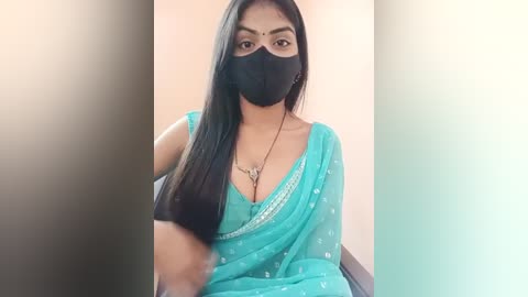 Media: Video of a South Asian woman with long black hair, fair skin, wearing a turquoise saree, black mask, and a necklace, sitting indoors against a blurred background.