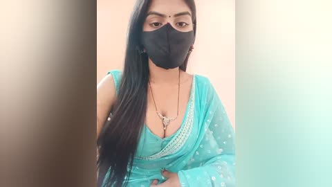 Media: Video of a South Asian woman with long black hair, wearing a teal saree with a low-cut neckline, black face mask, and gold jewelry, against a beige backdrop.